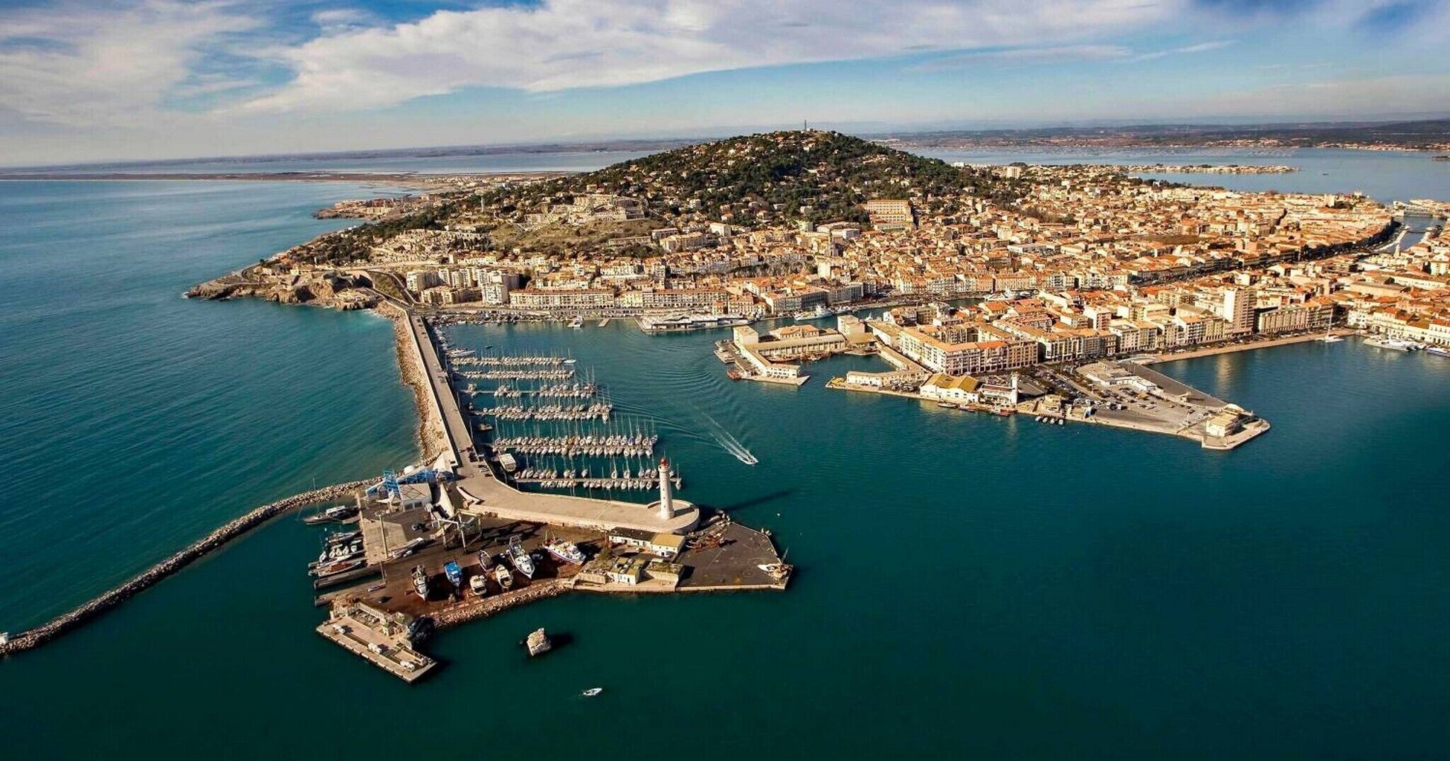 Vue aérienne d’une ville côtière, avec une colline imposante, une marina où sont amarrés de nombreux bateaux et une structure fortifiée.