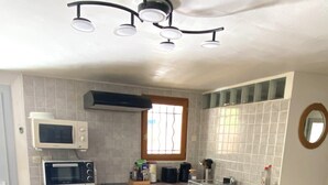 Een kleine keuken met een koelkast, magnetron en een broodroosteroven. Er is een raam met tralies, een spoelbak en een kleine prullenbak.