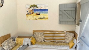 Een knus zithoekje met een comfortabele bank, een houten stoel en een tafel. Een schilderij van een strandbeeld siert de muur, en een bijzondere lichtarmatuur met gaatjes hangt erboven.
