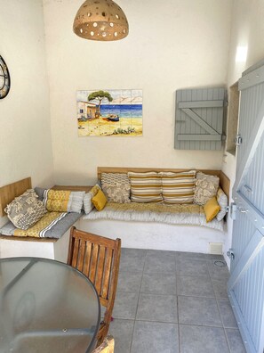 Un rincón acogedor con un banco acolchado, una silla de madera y una mesa. Un cuadro con una escena de playa decora la pared, y una lámpara original con diseño perforado cuelga arriba.