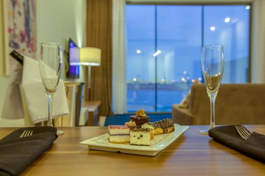 Mesa de jantar com um prato de sobremesas e duas taças de champanhe, em um quarto com vista para a paisagem urbana ao entardecer.