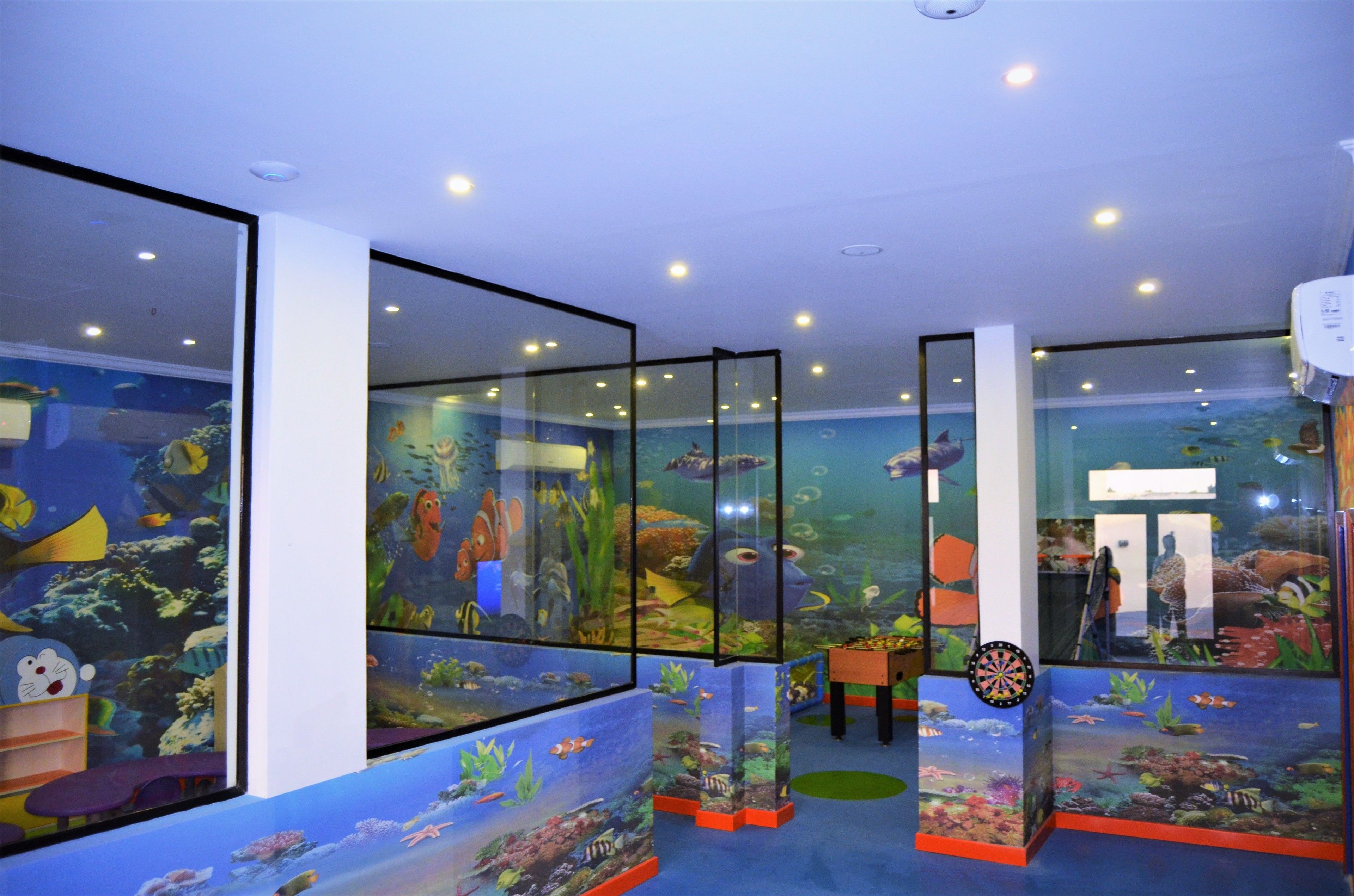 Un aquarium intérieur avec des murs en verre, abritant différentes espèces de la vie marine, et équipé d’un jeu de fléchettes.