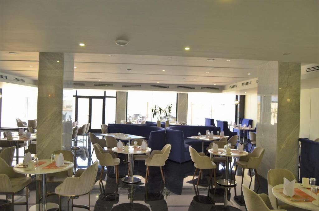 Un restaurante moderno con mesas y sillas redondas, un área de estar azul y ventanas amplias que dejan entrar la luz natural.