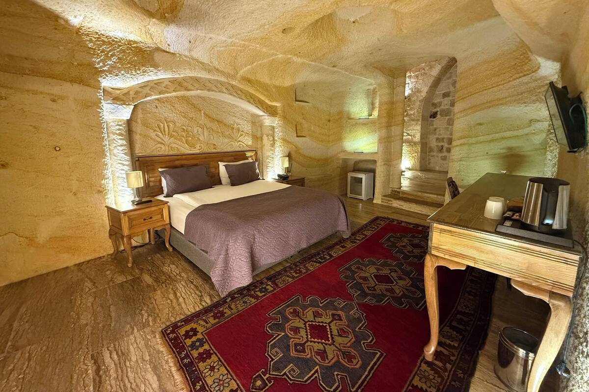 Una habitación con cama, mesitas de noche, escritorio, silla, alfombra y pared de piedra.