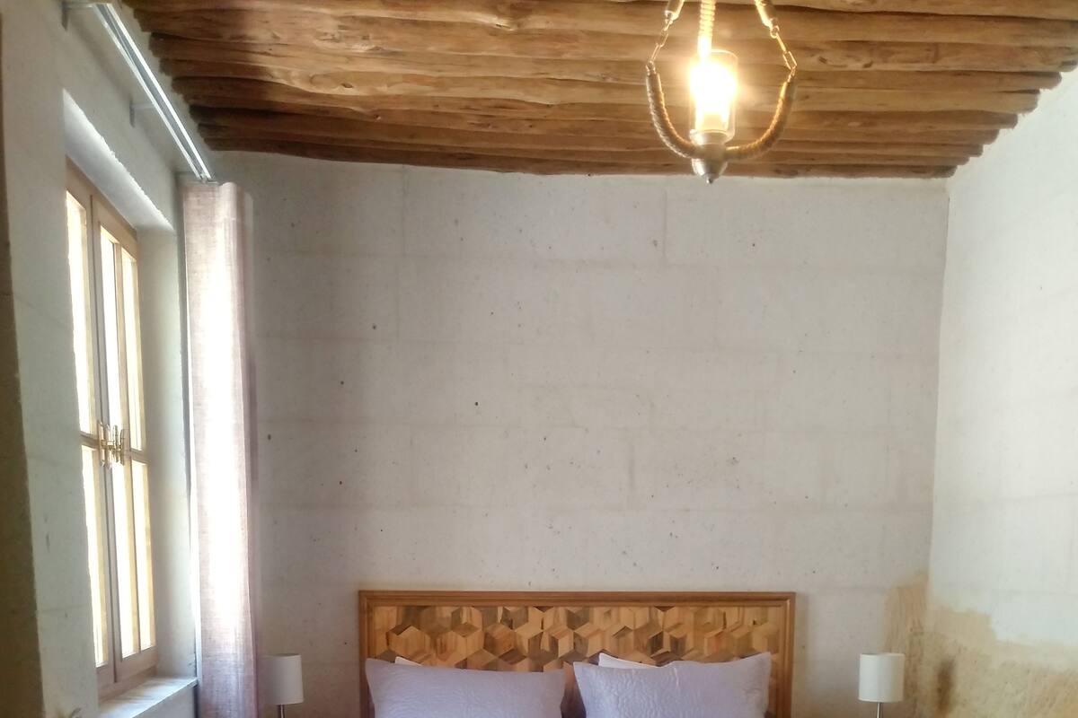 Un dormitorio con cabecera de madera, suelo de mármol, cama con ropa de cama blanca, dos mesitas de noche con lámparas y una ventana con barrotes.