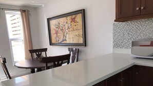 A kitchen with a white countertop, wooden cabinets, a dining table with chairs, and a framed artwork on the wall.