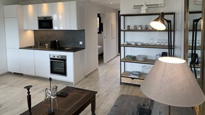 A modern kitchen with white cabinets, a black countertop, and a built-in oven. There is a dining area with a wooden table and a lamp. - New- Beach 2min, Direct accès marina, 2km from Saint-Tropez (Les marines de Cogolin)
