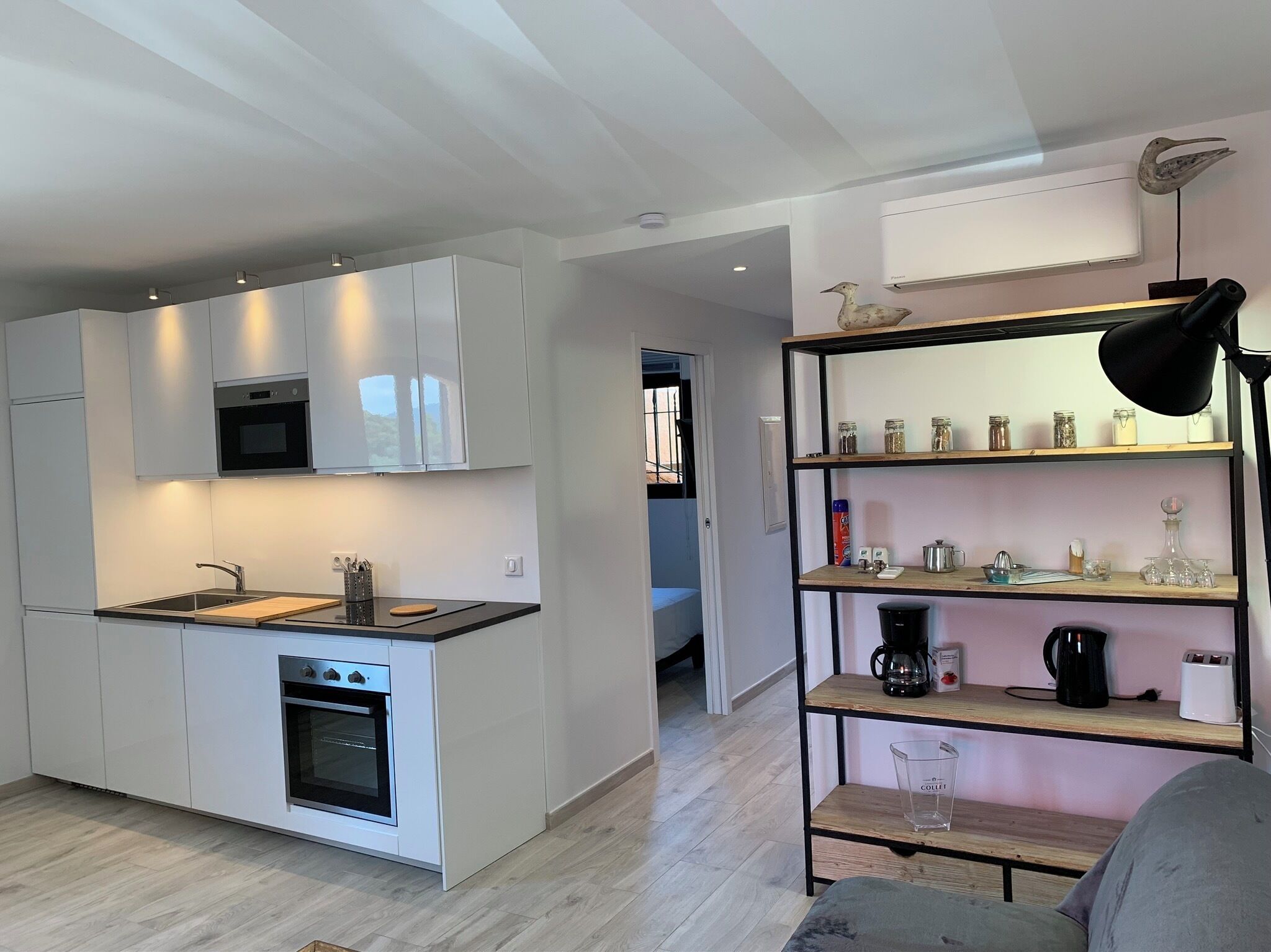 Eine moderne Küche mit weißen Schränken, einem eingebauten Backofen und einer Mikrowelle. Ein Holzregal beherbergt verschiedene Küchenutensilien, und eine schwarze Lampe sorgt für Licht.