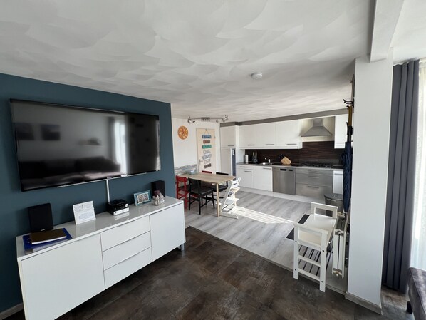 A modern living room with a flat-screen TV, a white entertainment unit, and a dining area with a wooden table and chairs.