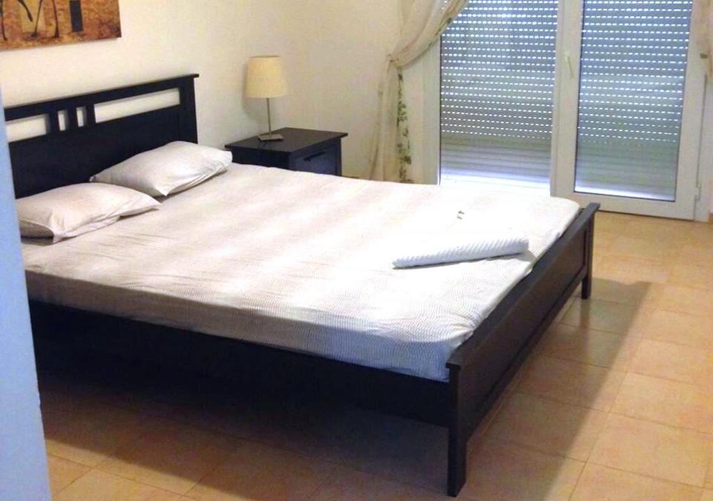 A neatly made bed with a white comforter and pillows, a small bedside table with a lamp, and a window with blinds.