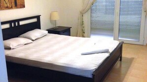 Ein ordentlich bezogenes Bett mit weißer Tagesdecke und Kissen, ein kleiner Nachttisch mit Lampe und ein Fenster mit Jalousien.