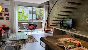 A modern living room with a glass dining table, a black chair with a red cushion, a TV on a wooden stand, and a magazine on the table. - LUXURY DUPLEX, fully equipped, 2Bd, pool & terrace (Palermo)