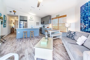A modern kitchen with grey cabinets, a central island with stools, a dining table, and a grey sofa with patterned cushions.