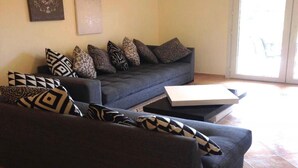 Een woonkamer met een donkergrijze bank, gekleurde kussens, een witte salontafel en een deur die naar buiten leidt.
