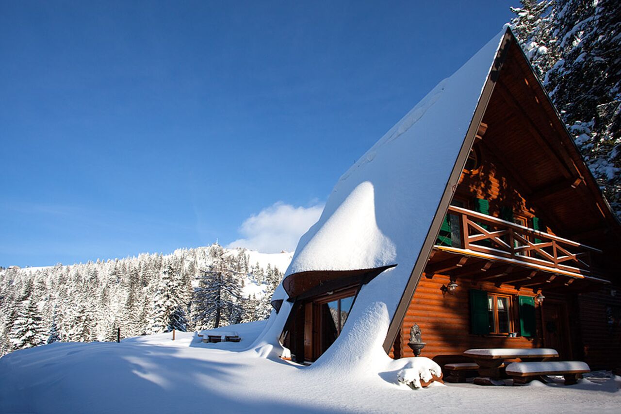 Ein mit Schnee bedecktes Blockhaus mit geneigtem Dach, Holzfassade und einem großen Fenster.