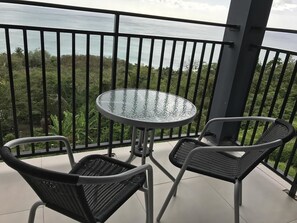 Varanda com mesa redonda de vidro e duas cadeiras de vime, com vista para uma paisagem verdejante e o mar.