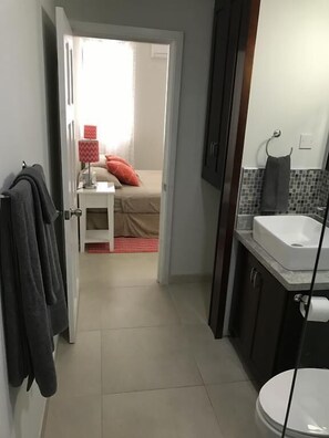 Corredor estreito que dá para um quarto com cama, mesa de cabeceira e abajur. Um banheiro com pia e vaso sanitário é visível à direita.