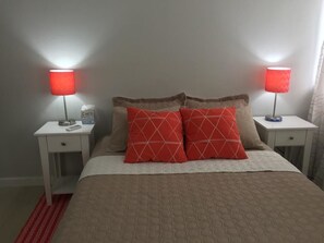 Uma cama com um edredom bege, duas almofadas vermelhas com padrão geométrico e duas luminárias de cabeceira com abajures vermelhos.