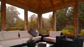 A covered outdoor seating area with a white sofa, a black wicker chair, and a glass coffee table. - Chalet Sean, peaceful retreat on beautiful mountain brook and walk to river.. (Sutton)