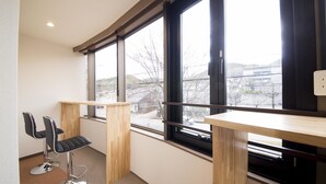 Una habitación moderna con dos mesas de madera y dos sillas negras, ventanas amplias con vistas a edificios y árboles, y una alfombra de color claro.
