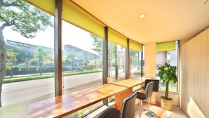 Una habitación moderna con un escritorio de madera y dos sillas, ventanas amplias con vista a una calle y edificios, y una planta en maceta.