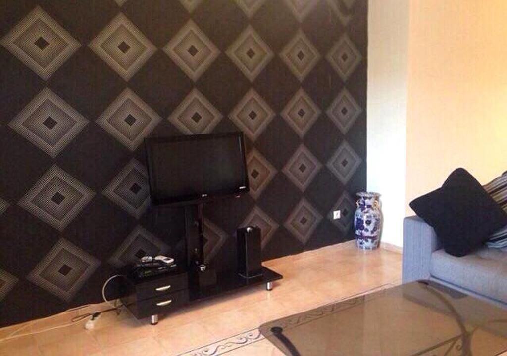 A living room with a flat-screen TV on a stand, a small table with a vase, and a sofa with a cushion.