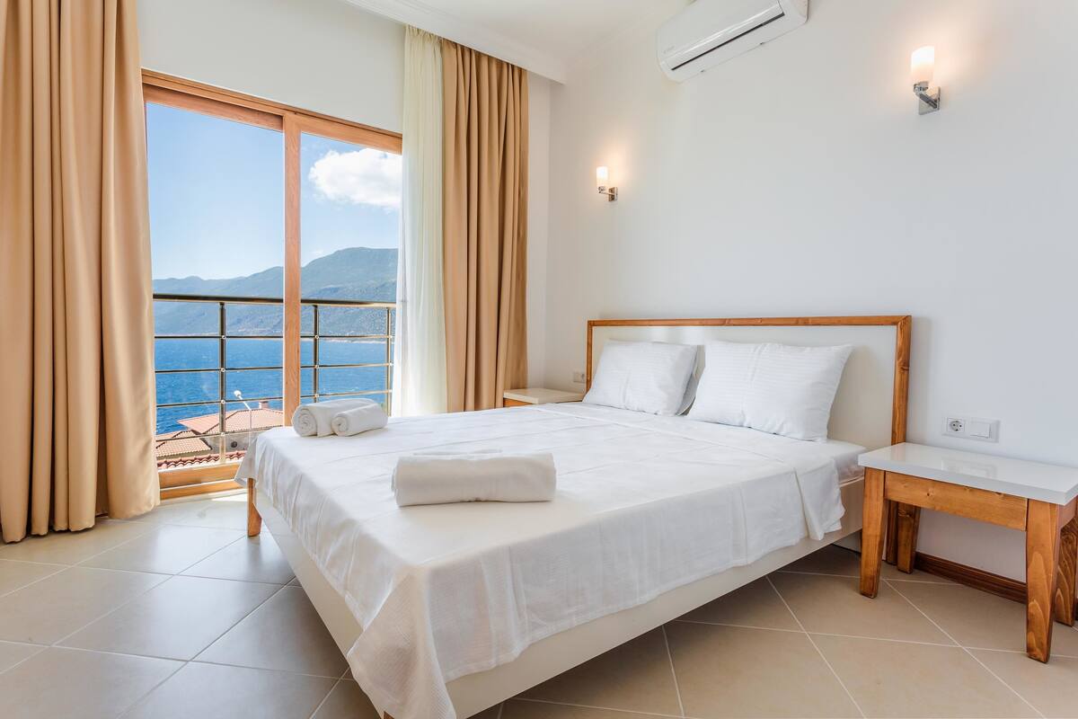 A hotel room with a large bed, a wooden nightstand, a window with curtains, and a view of the sea.