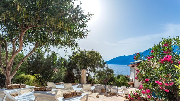Un espace extérieur ensoleillé, aménagé avec des chaises longues blanches et des marches en pierre menant à un bâtiment d’où l’on peut admirer la mer et les montagnes.