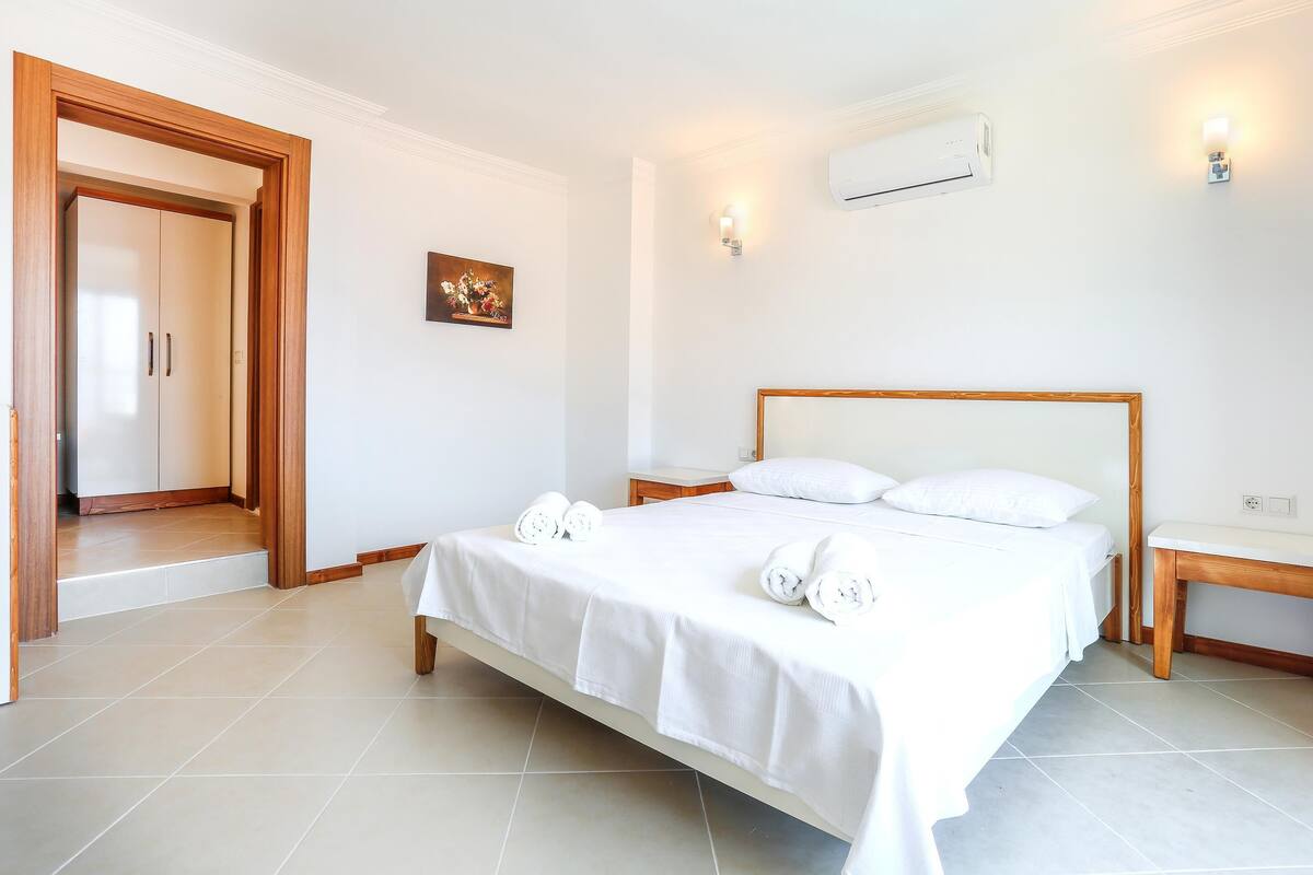 A hotel room with a bed, a bedside table, a wall-mounted air conditioner, and a painting on the wall.