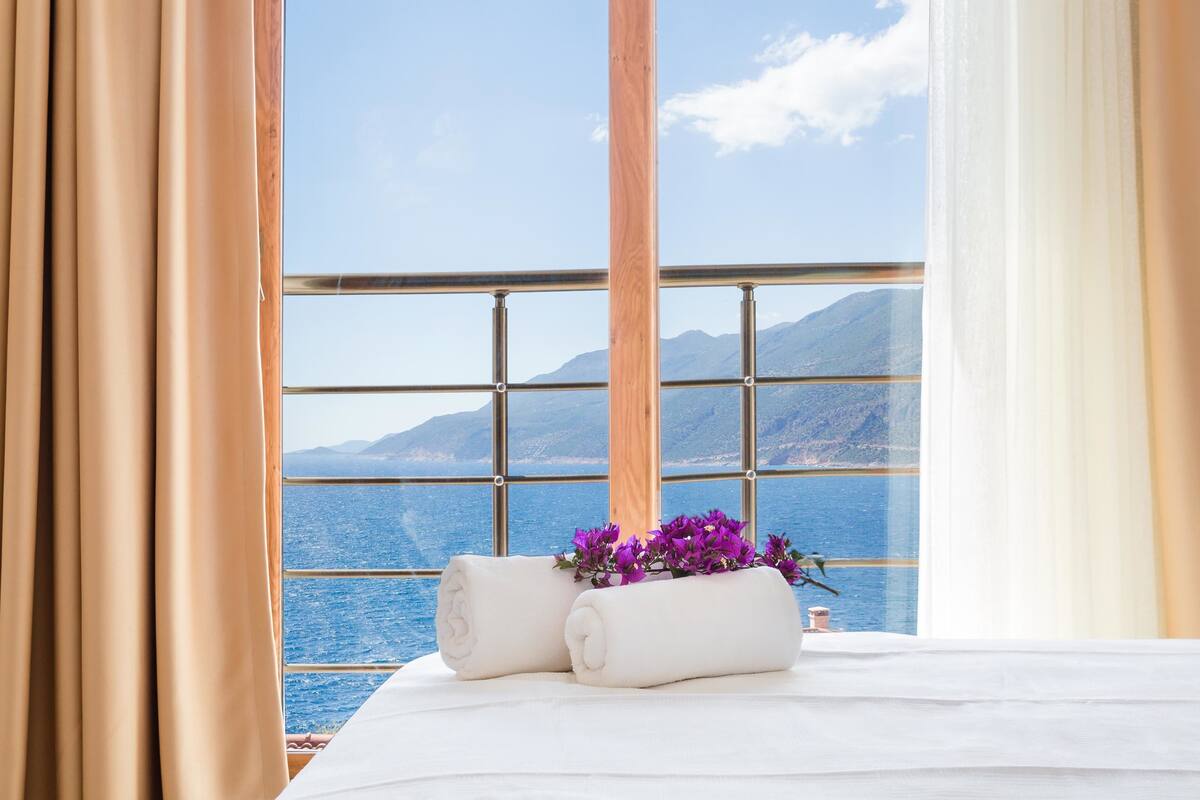 A bed with rolled towels and purple flowers, a window with a view of the sea and mountains, and beige curtains.