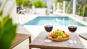 A table with a fruit platter and two glasses of red wine, a pool, and a blurred background of greenery and buildings. - Maison W (Kotor)