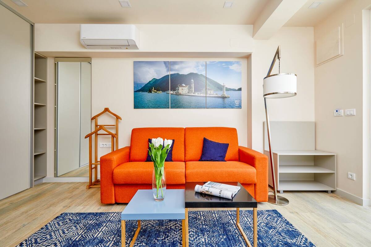 Una sala de estar con un sofá naranja, una mesa de centro azul, un jarrón con flores, un cuadro de una escena costera y una lámpara de pie.