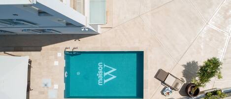 Vista aérea de una piscina con el logo 'Maison', rodeada por una zona pavimentada con un pequeño espacio de estar y plantas en macetas.
