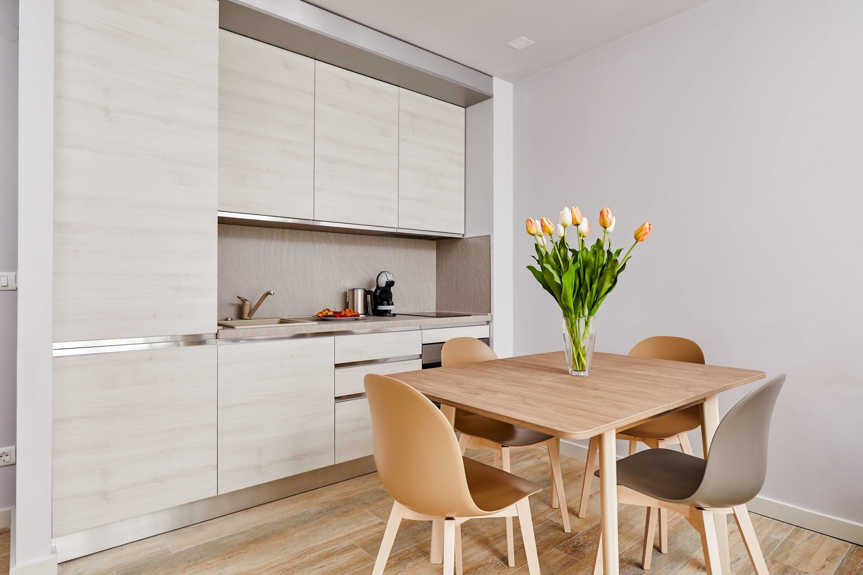 A modern kitchen with light wood cabinetry, a wooden dining table with four chairs, and a vase of tulips on the table.