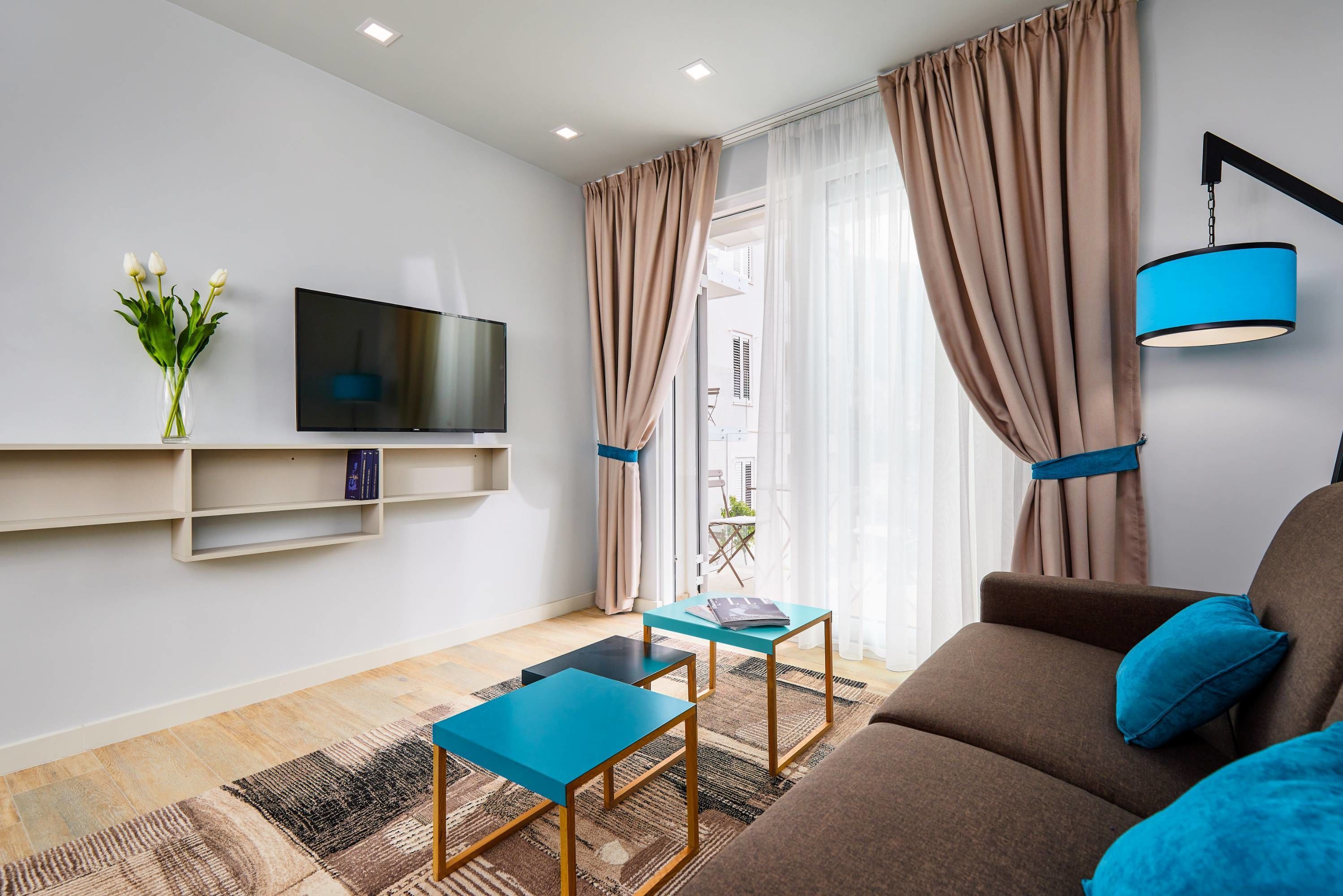 Una sala de estar moderna con un televisor de pantalla plana, dos mesas azules, un sofá marrón y una lámpara colgante azul.