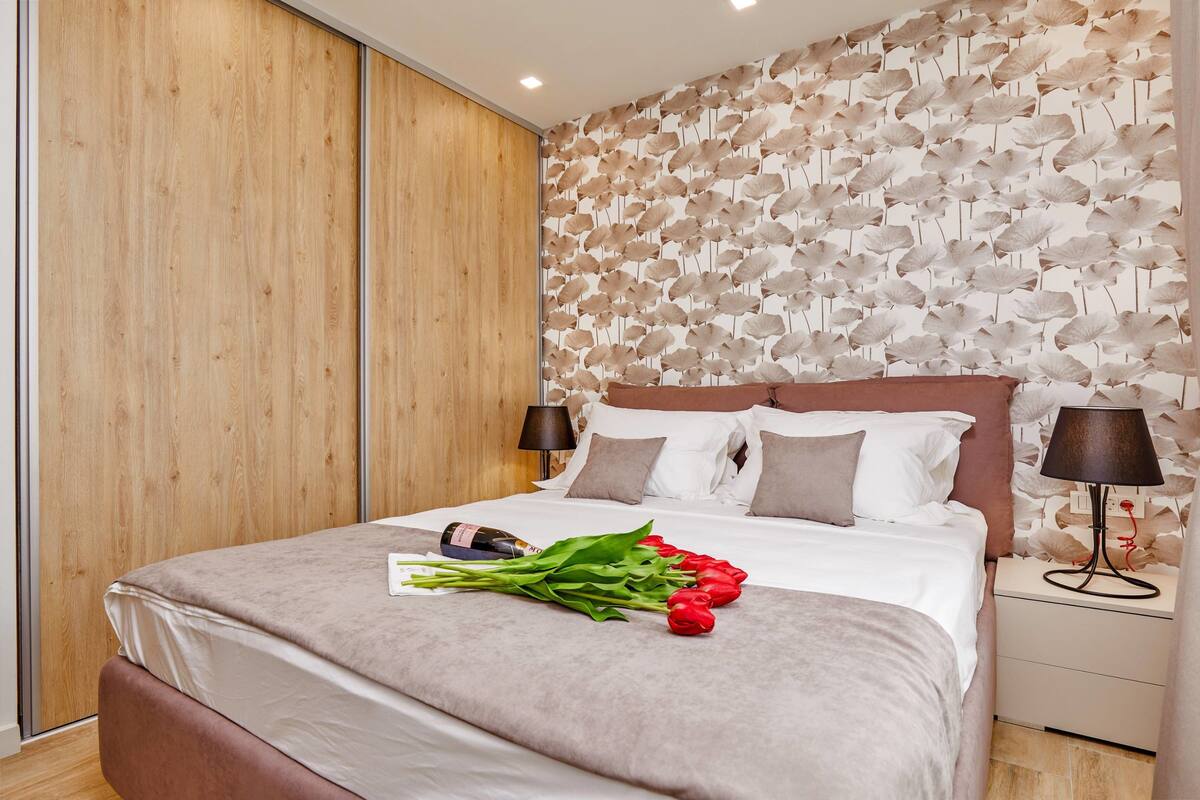 Una cama bien hecha con cubrecama gris, una mesita de noche con lámpara y una pared con diseño de hojas.
