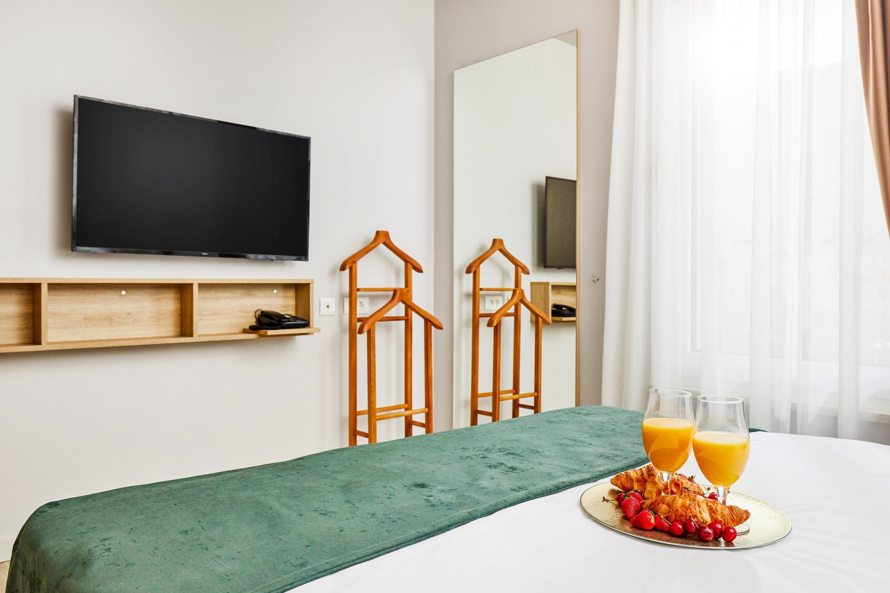 A hotel room with a bed, a flat-screen TV mounted on the wall, wooden shelves, a mirror, and a breakfast tray with croissants and juice glasses.