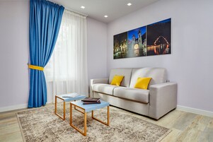 Una sala de estar moderna con un sofá, dos mesitas auxiliares, un tapete y un ventanal con cortinas azules.