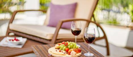 Une table en osier, sur laquelle sont disposés un plateau de fruits et deux verres de vin rouge, est placée sur une terrasse extérieure, avec un fauteuil de jardin en arrière-plan.