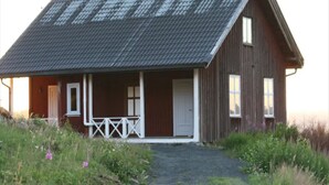 A wooden cabin with a sloped roof, a porch with a railing, and a gravel path leading to the entrance.