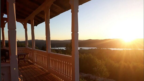A covered outdoor area with a wooden deck, white balustrades, and a view of a lake and hills during sunset.