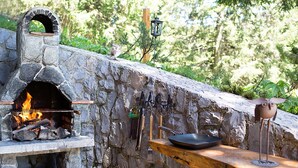 A stone-built outdoor kitchen with a wood-fired oven, a wooden counter, and cooking utensils.