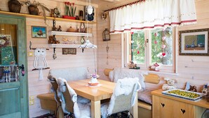 A cozy cabin interior with a wooden dining table, cushioned chairs, a bench, and a small kitchen area with a tray of food.
