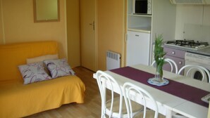 A compact living space with a yellow sofa, a dining table with chairs, a microwave, a refrigerator, and a stove.