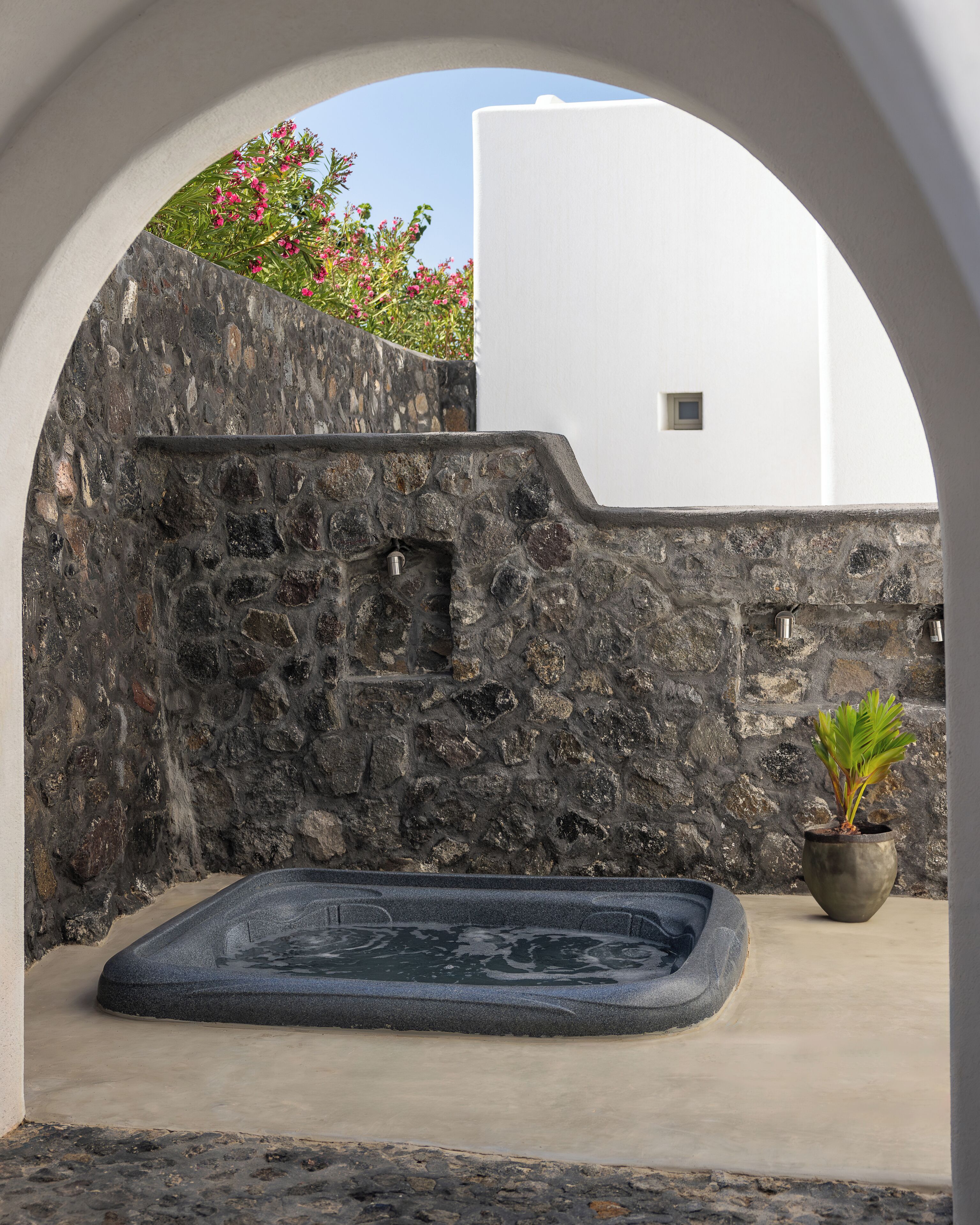 A stone wall with a small potted plant, a stone bench, and a white building in the background.
