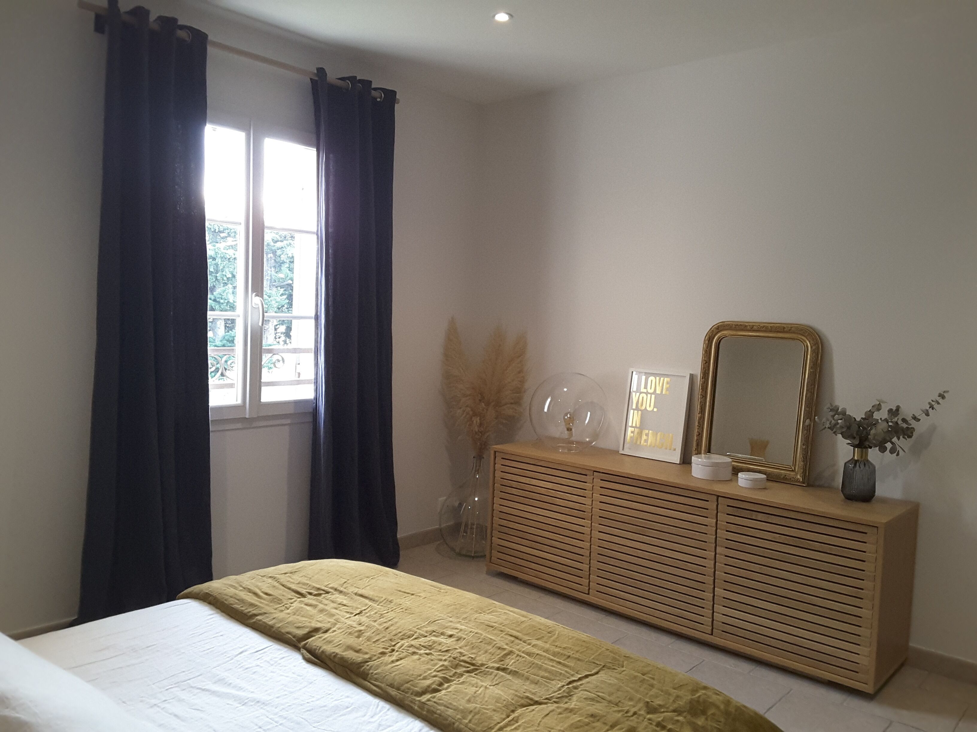 Een slaapkamer met een bed, een houten dressoir, een spiegel en een raam met gordijnen.