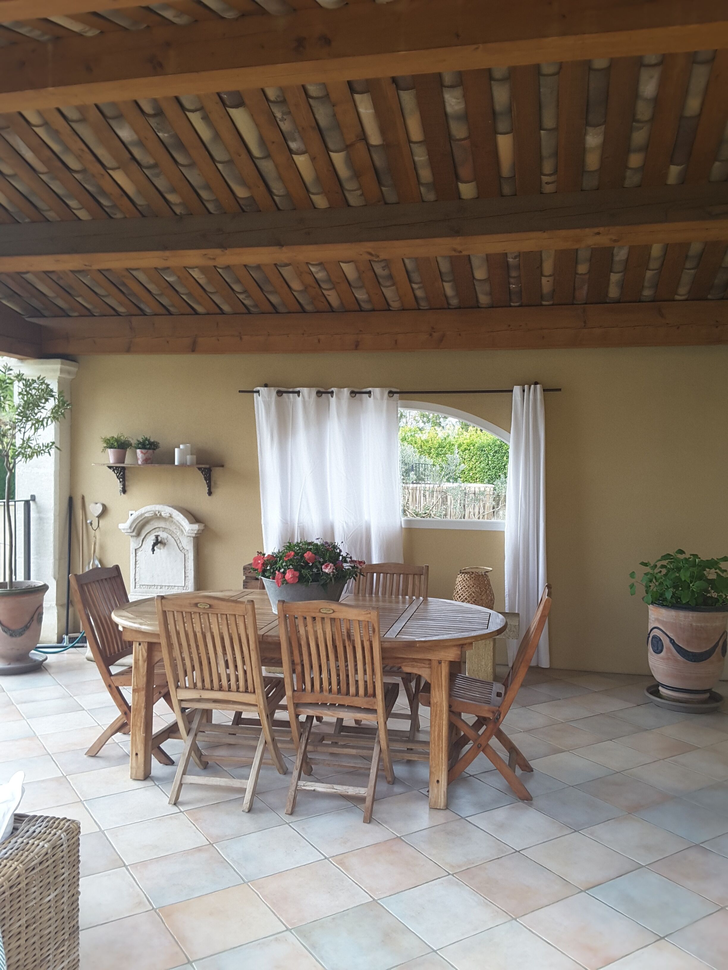 Een overdekt terras met een houten tafel en stoelen, potplanten en een raam met gordijnen.