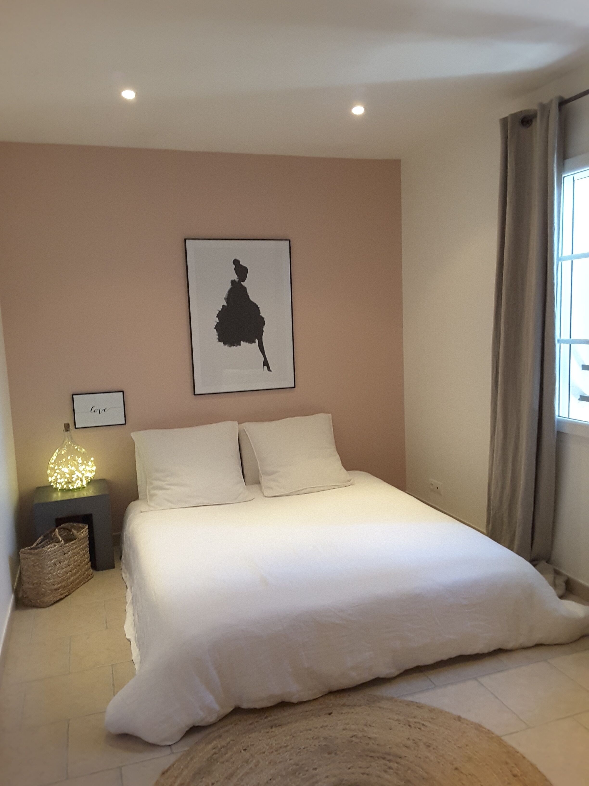 Ein Schlafzimmer mit einem großen Bett, einem Wandbild, einem Nachttisch mit Lampe und einem Fenster mit Vorhängen.