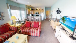 A living room with striped sofas, a TV, and a kitchen in the background. - Escape to River Creek | The Perfect NC Golf Condo | RC 1602 (Sunset Beach)