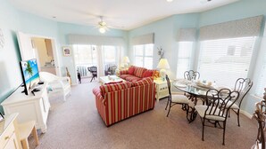 A living room with a sofa, a dining table with chairs, a TV, and a ceiling fan. - Escape to River Creek | The Perfect NC Golf Condo | RC 1602 (Sunset Beach)